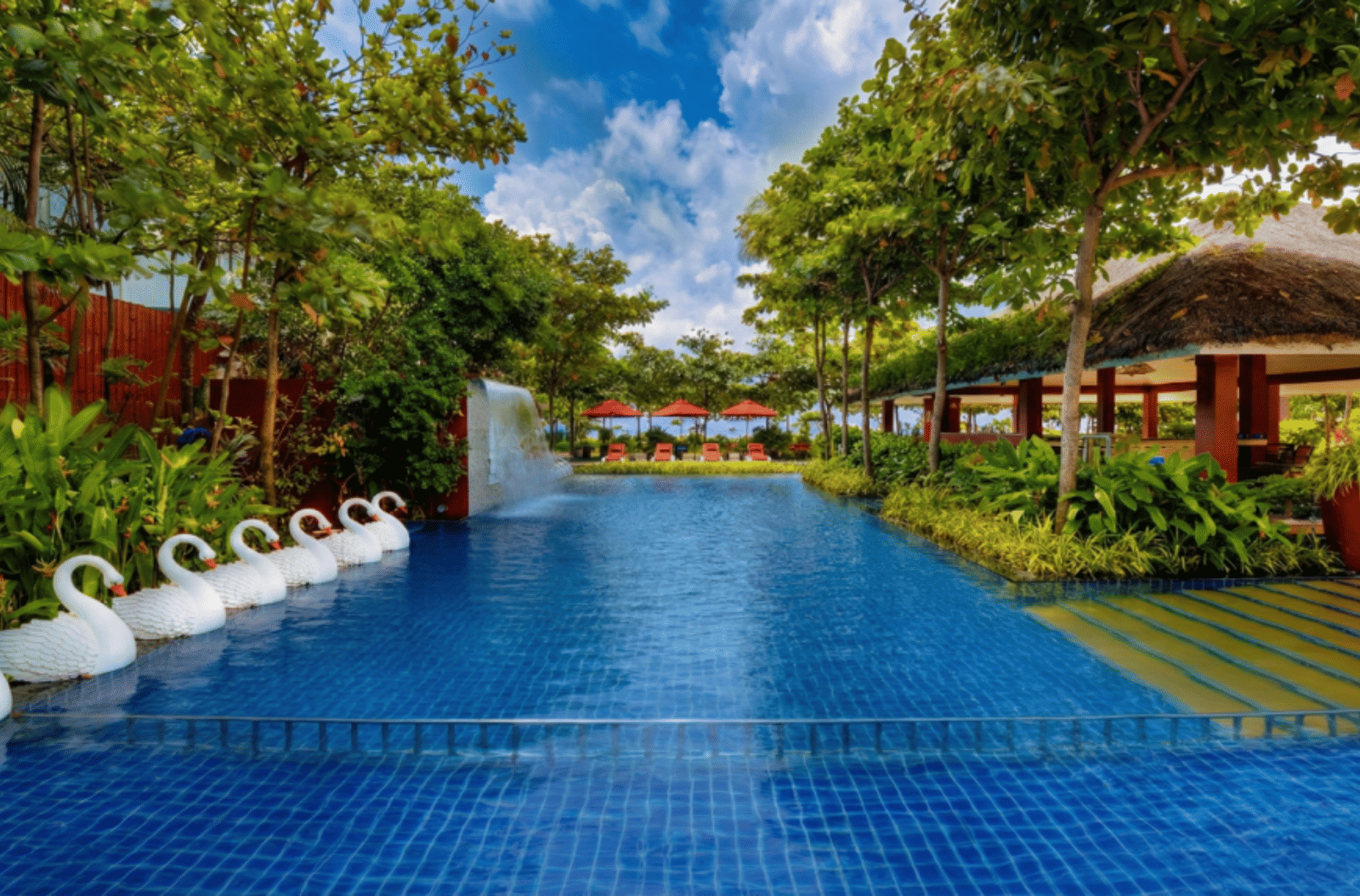 long, narrow outdoor swimming pool with blue tiles, surrounded by lush green trees, featuring a small waterfall on the left and a row of white swan statues near the edge.