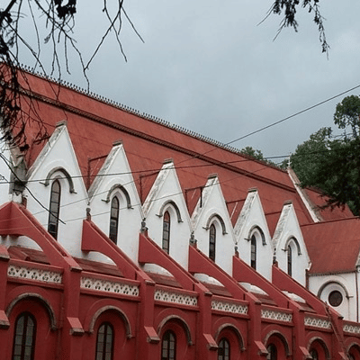 A gothic-style church with tall spires and arched windows, nestled amidst trees under a cloudy sky.