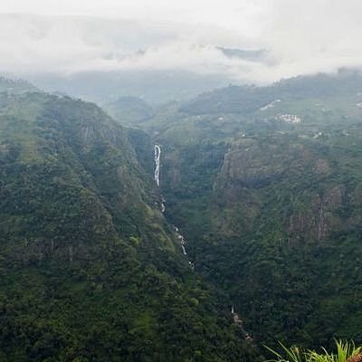 A view of a large waterfall cascading down a deep valley, surrounded by dense forest and mist-covered hills.