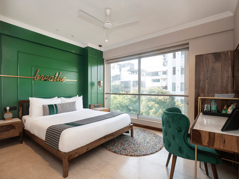 A bedroom at Theory9 - Premium Service Apartments, Bandra, featuring a large bed with white linens and a green accent wall with the word 'breathe' in gold, a wooden nightstand, a desk with a green velvet chair, and a large window overlooking trees.