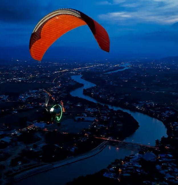 A paraglider flying above a river and city lights during twilight, capturing breathtaking night-time views near Alibu Resort.