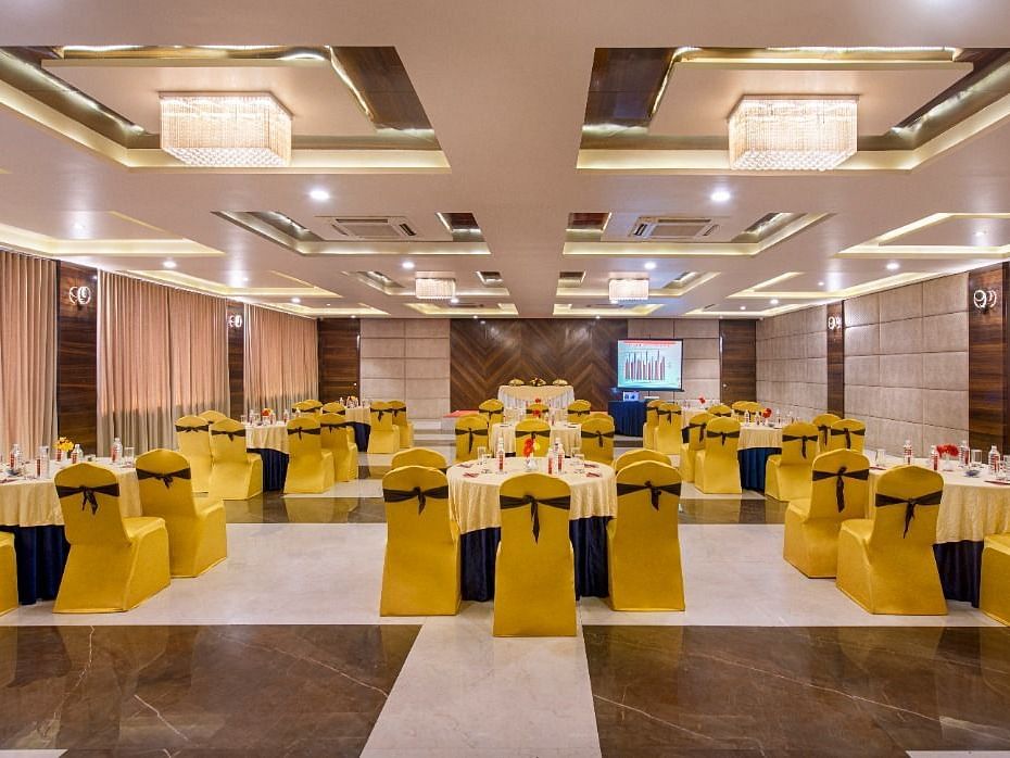 A spacious event hall at Lords Inn Porbandar, is prepared with round tables and chairs with yellow sashes, ready for guests.