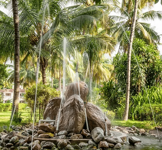 Stone fountain surrounded by lush greenery, palm trees, and trimmed bushes in a garden setting with water gently spraying upward