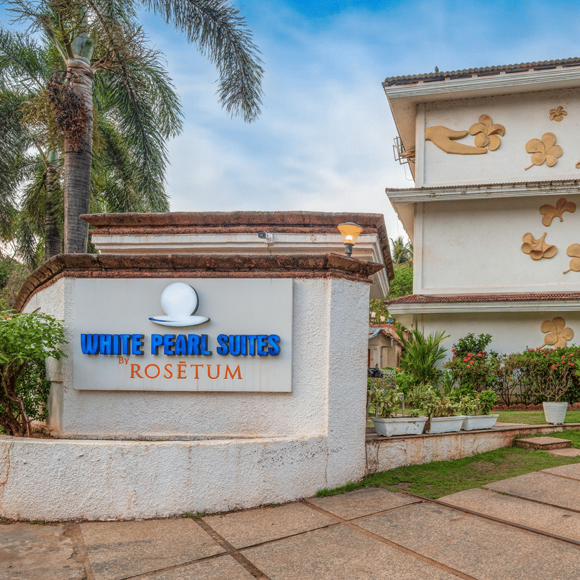 Entrance sign and facade of White Pearl Suites by Rosetum Hotels in Anjuna, Goa, surrounded by tropical palms and floral wall art.