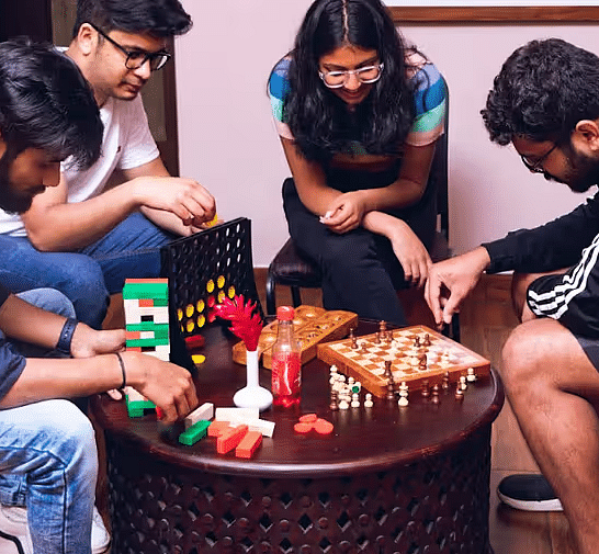 Four friends playing fun indoor games like Jenga, chess, Connect Four, and other board games around a round wooden table with snacks.