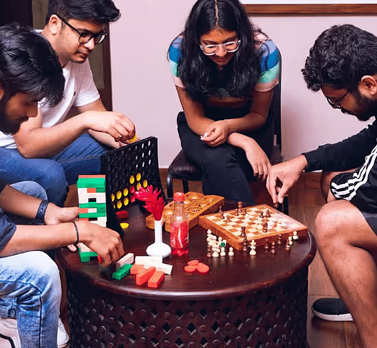 Four friends playing fun indoor games like Jenga, chess, Connect Four, and other board games around a round wooden table with snacks.