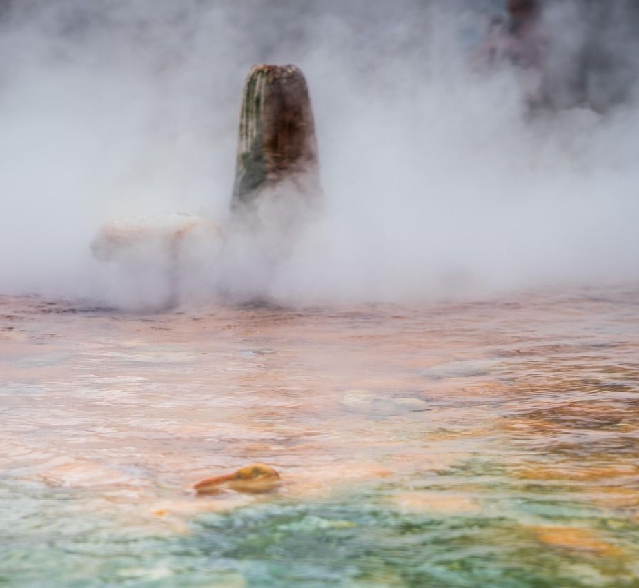 A wide, steaming geothermal hot spring with colourful mineral-stained water and a tall, chimney-like rock formation emerging from the mist.