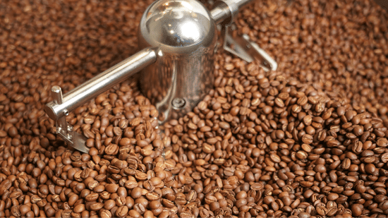 A close-up view of freshly roasted coffee beans spread across a roasting surface at Unseen Boutique Hotel Assagao Goa with a metal stirring tool visible among the beans.
