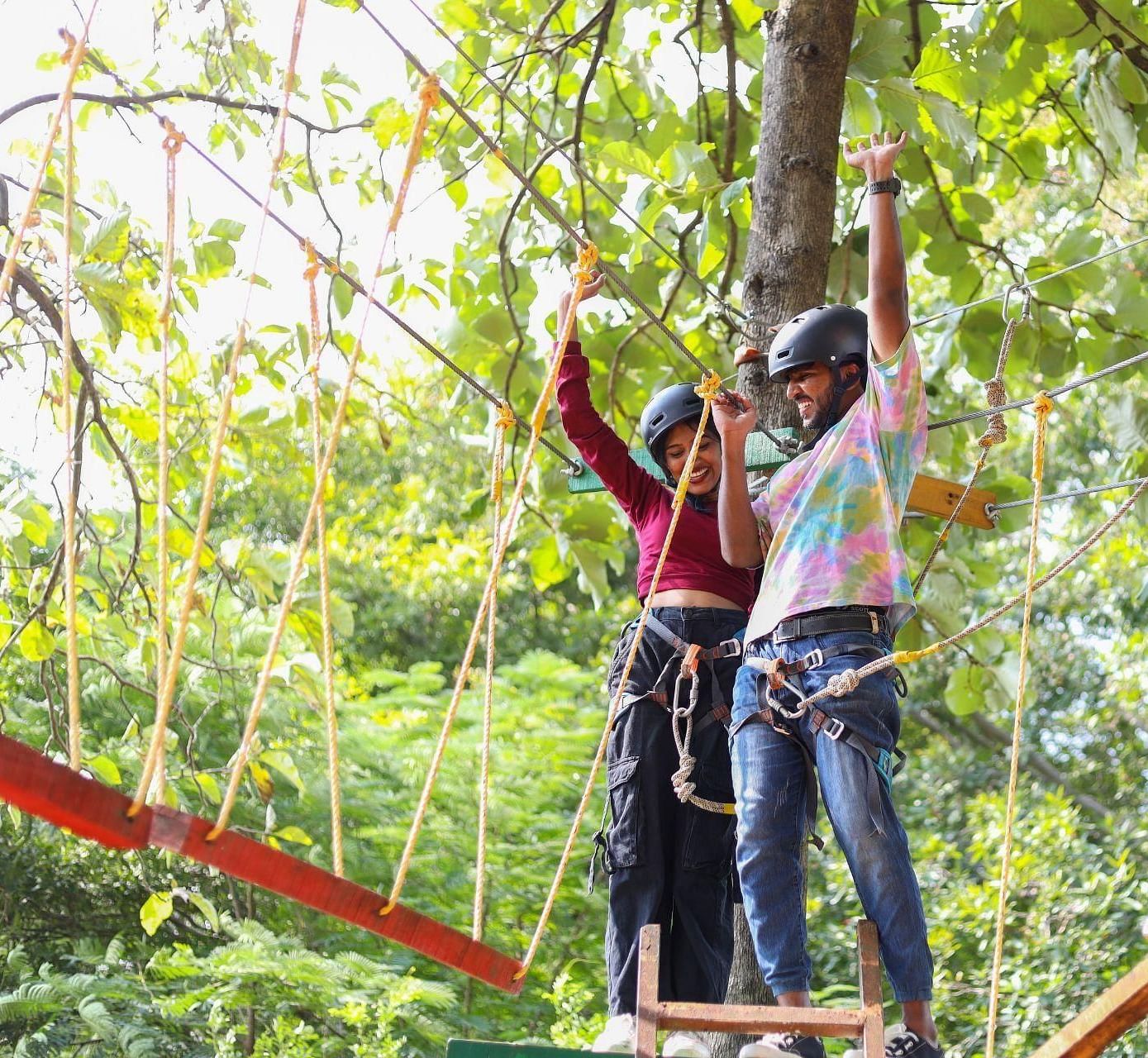 Two individuals on a ropes course amidst trees, harnessed and smiling, enjoying an adventure activity at Avana Resort.