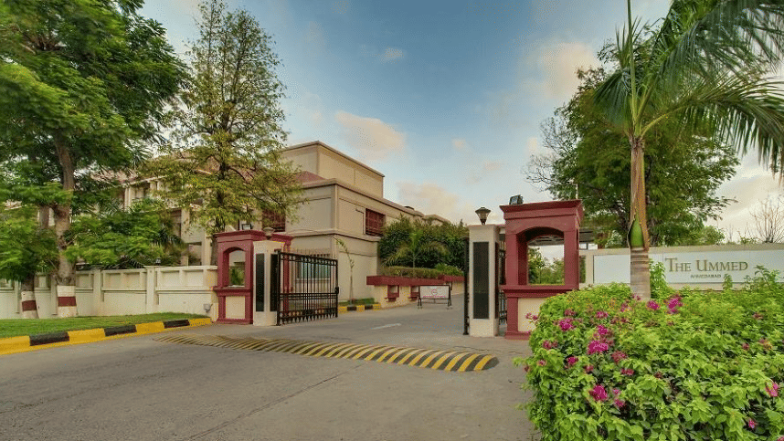 The entrance of The Ummed, Ahemdabad showcasing a gate, boundary wall, lush greenery under a partly cloudy sky, and a welcoming driveway.