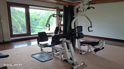 Hotel fitness room at Vedic Village Spa Resort with multi-functional gym equipment, exercise machines, and large windows overlooking greenery.
