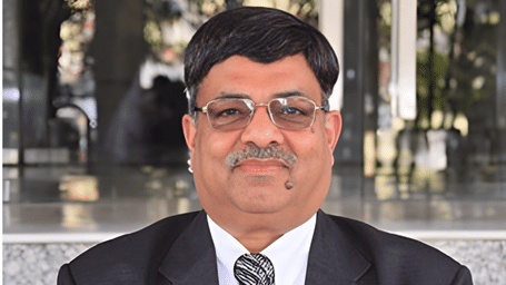 Portrait of a middle-aged Indian man with a mustache and glasses, wearing a black suit and a patterned tie.