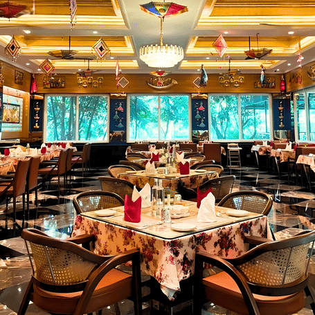 Rangmahal restaurant at MAYFAIR Tea Resort, Siliguri features tables and chairs arranged across a floor. The ceiling contains recessed panels with lighting fixtures and decorations that hang above the tables. 