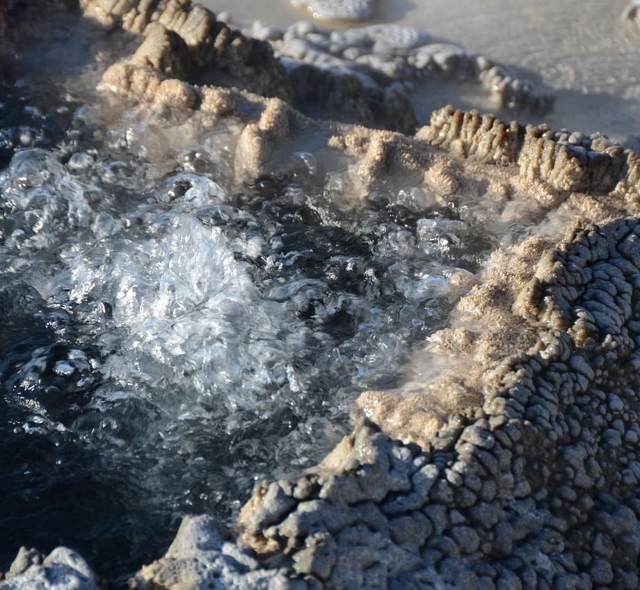 A close-up shot of boiling water bubbling vigorously within a geothermal pool bordered by rough, grey mineral deposits.