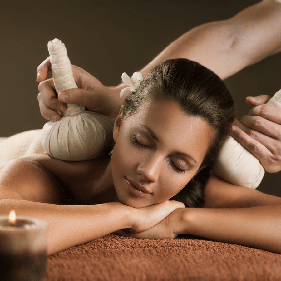 A woman relaxing with her eyes closed as a therapist applies herbal compresses to her shoulders, with lit candles in the foreground