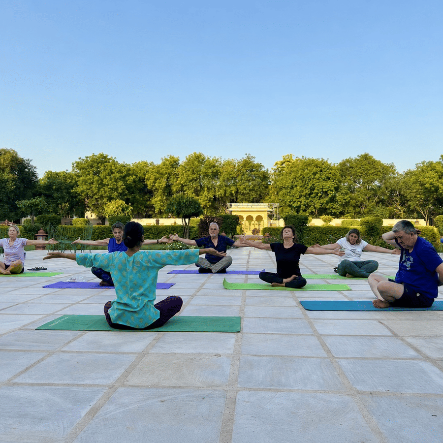 A group of people practicing yoga on colorful mats during an outdoor session on a paved courtyard surrounded by lush green trees.