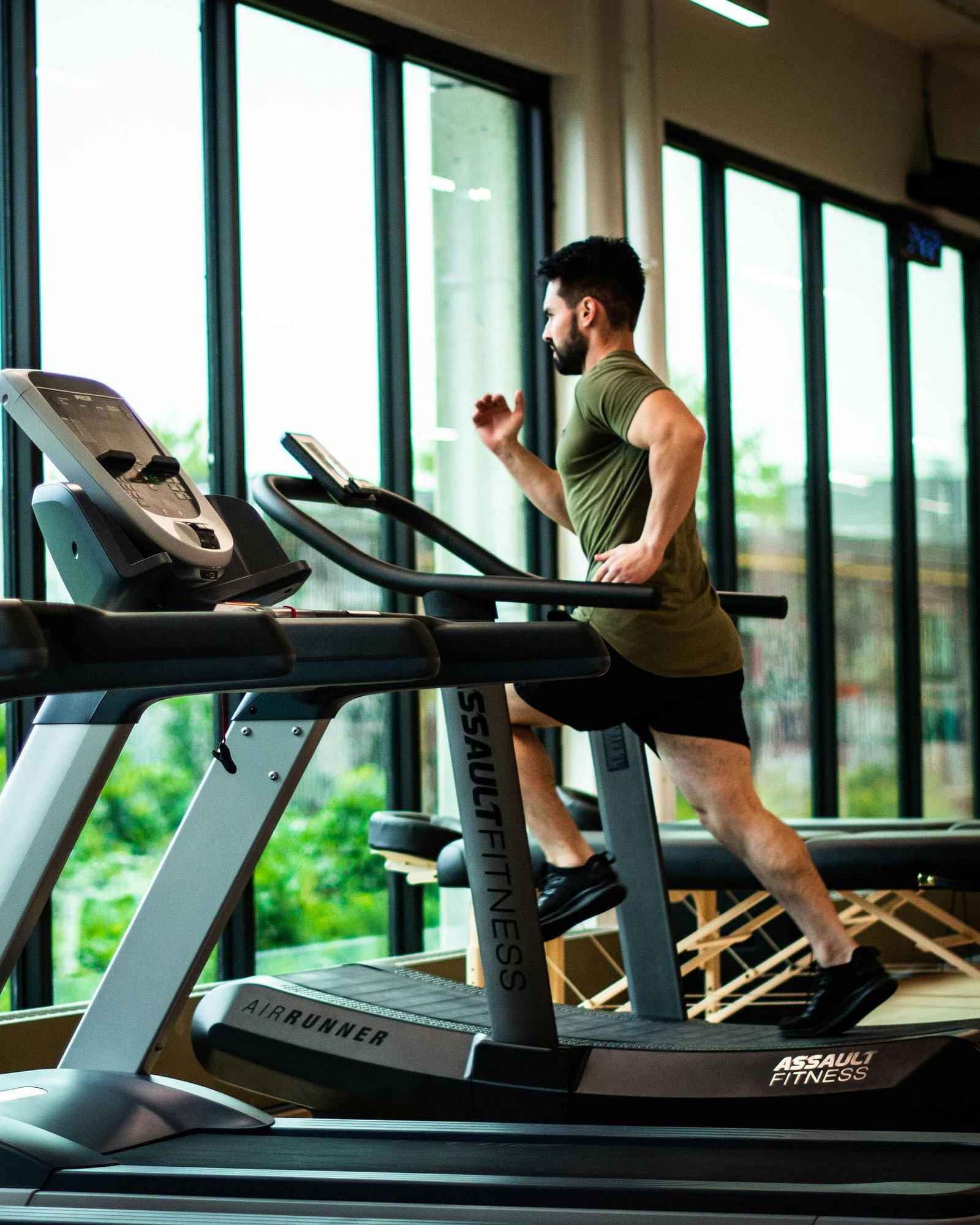 A person exercises on a treadmill in a well-lit gym with large windows overlooking greenery.