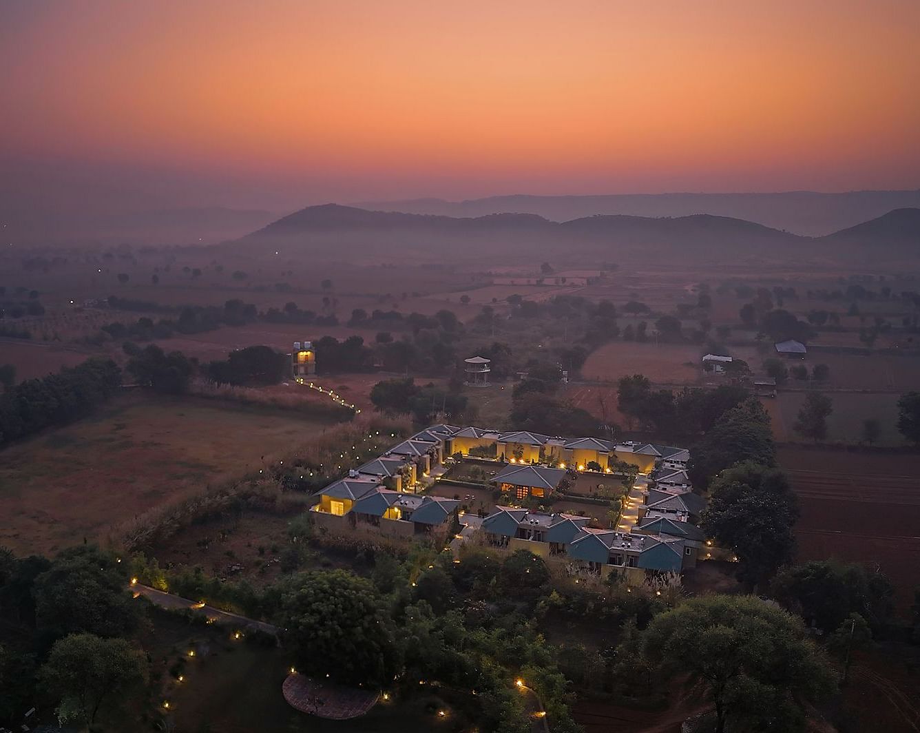 An aerial shot of a symmetrically arranged resort with illuminated villas, surrounded by trees and a hazy landscape at sunset - Singhvilas, Ranthambore