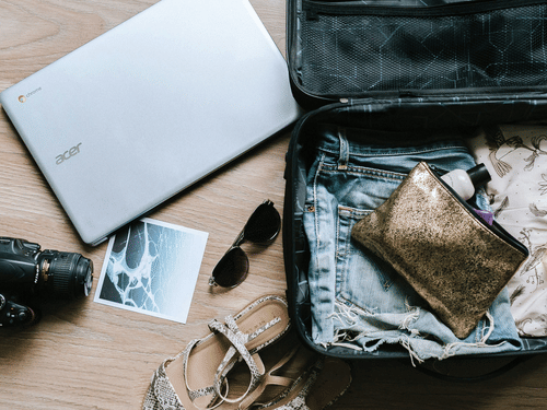 An open suitcase with neatly packed clothes, laptop, camera, and travel essentials laid out on a wooden floor, ready for a trip.