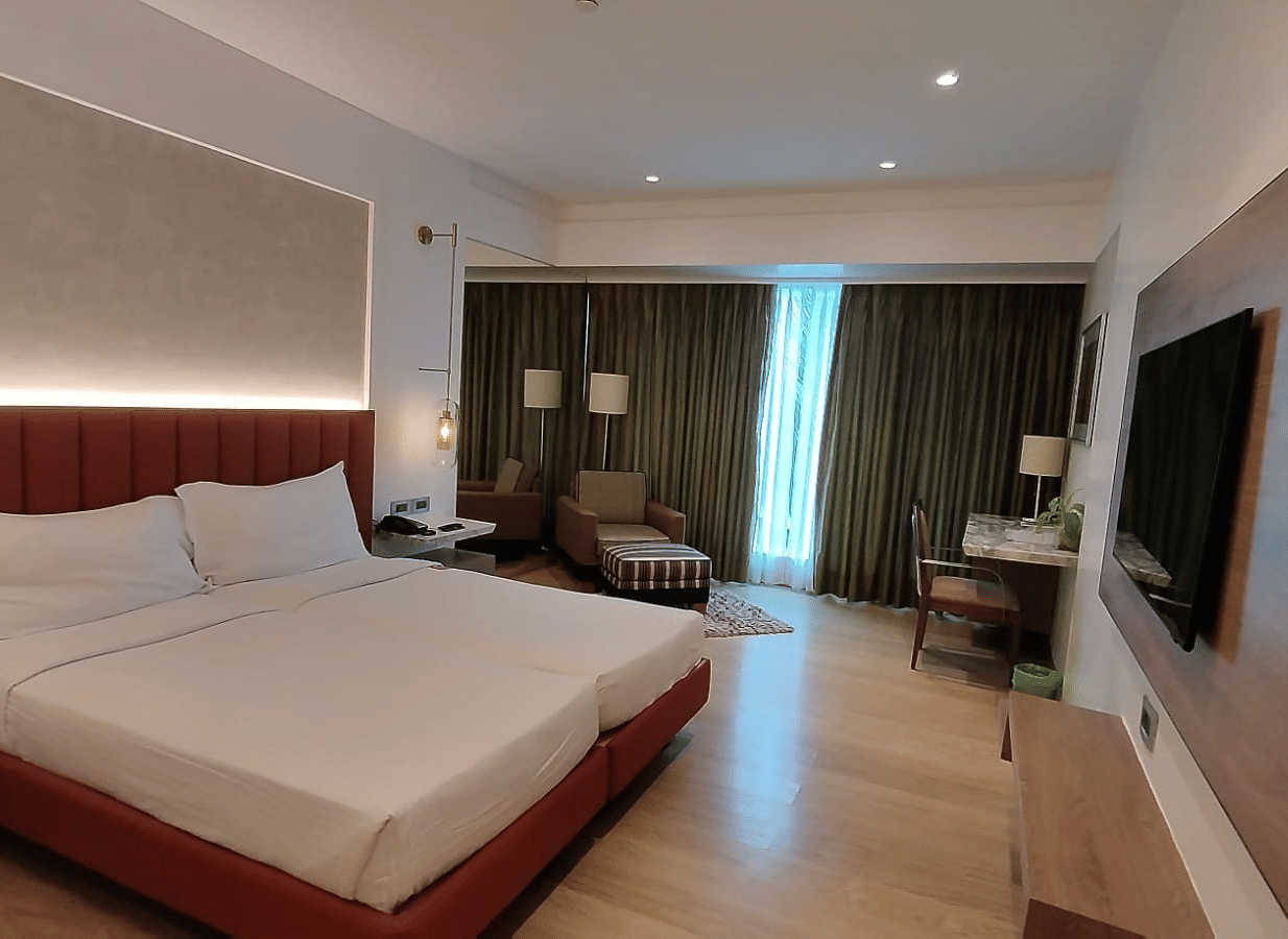 A well-lit executive bedroom in Mirage Hotel, featuring twin beds, decorative head-rest, lounge chairs, a wooden panelled entertainment unit and a large wall to wall window with closed drapes.