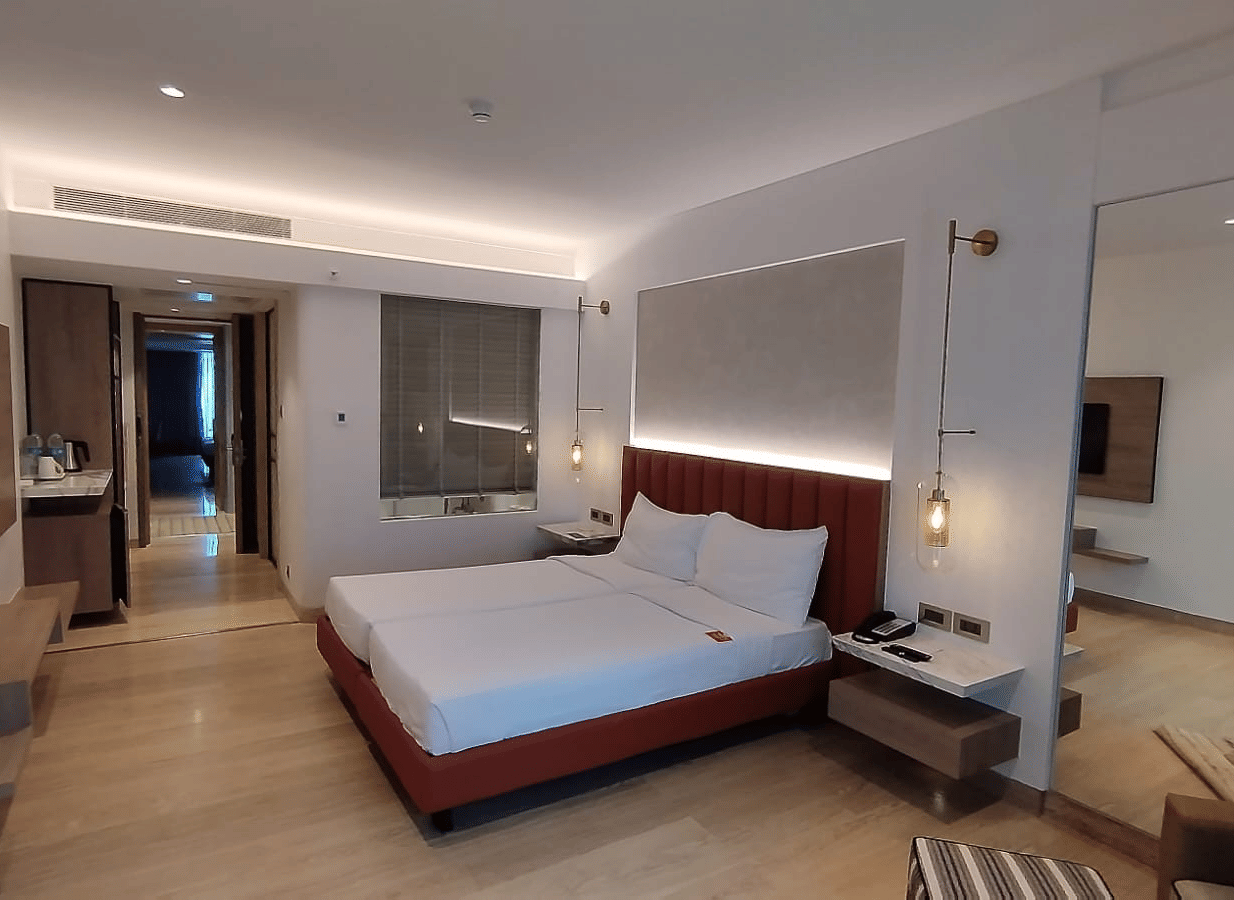 A warmly lit executive bedroom in Mirage Hotel, featuring twin beds, a large mirror, lounge chairs, and an entertainment unit.