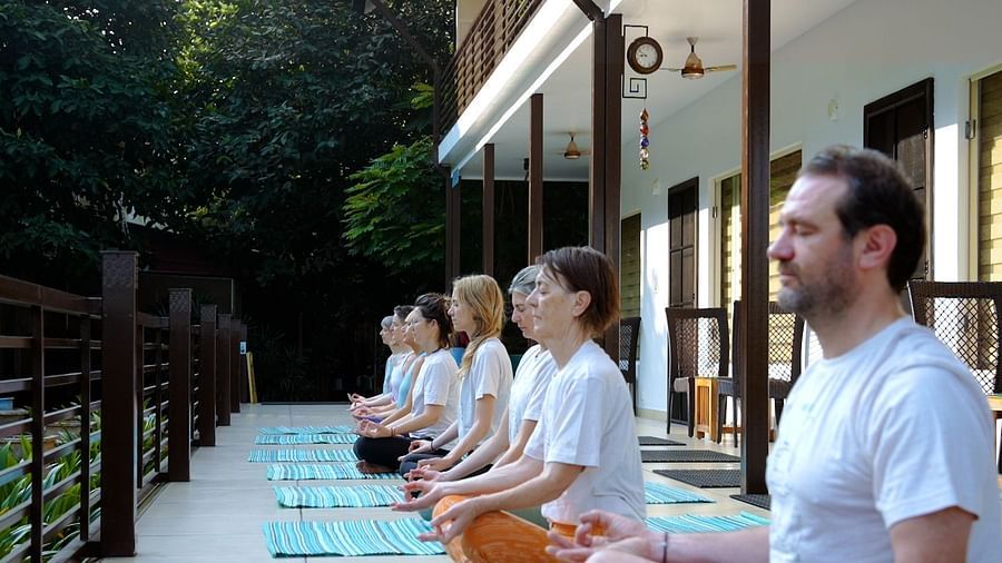 A group of people sit in a row on yoga mats, practising meditation or yoga outdoors on a patio.