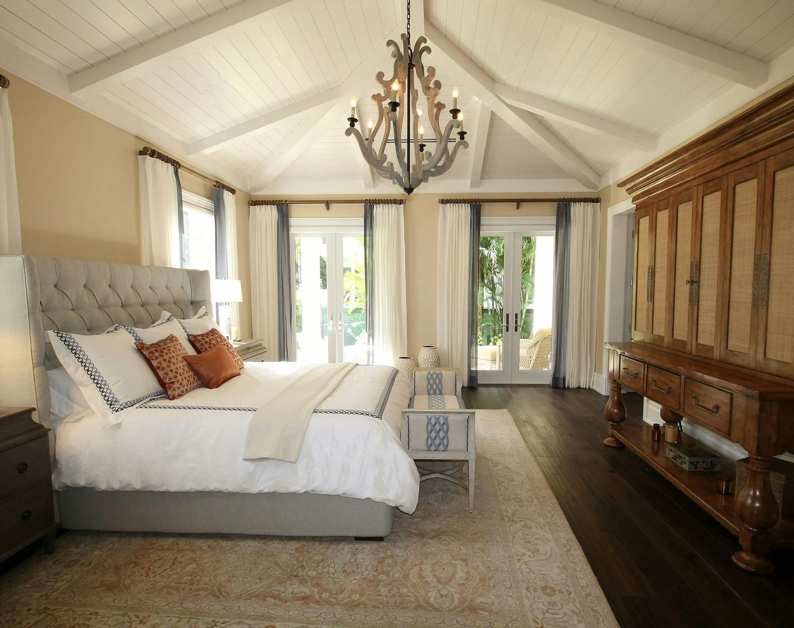 A spacious bedroom with a white bed, vaulted ceiling, chandelier, and a large wooden wardrobe.