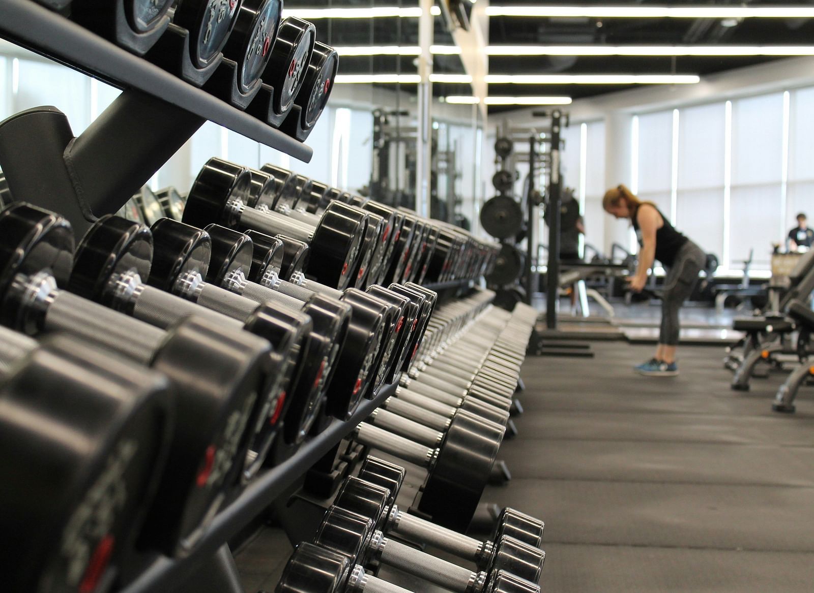 An image of a gym with dumbbells in front and a woman in the background