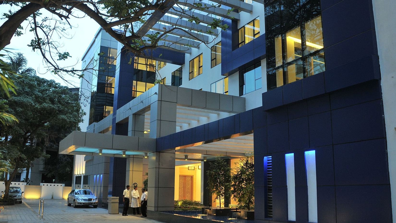 The exterior of a modern, multi-story building with blue and grey accents, illuminated at dusk - Adarsh Hamilton.