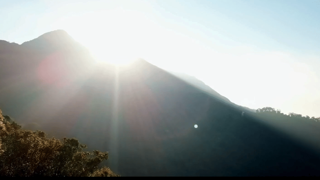 Sun rising over the misty green mountains of Munnar, Kerala, with a bright lens flare over the peaks.