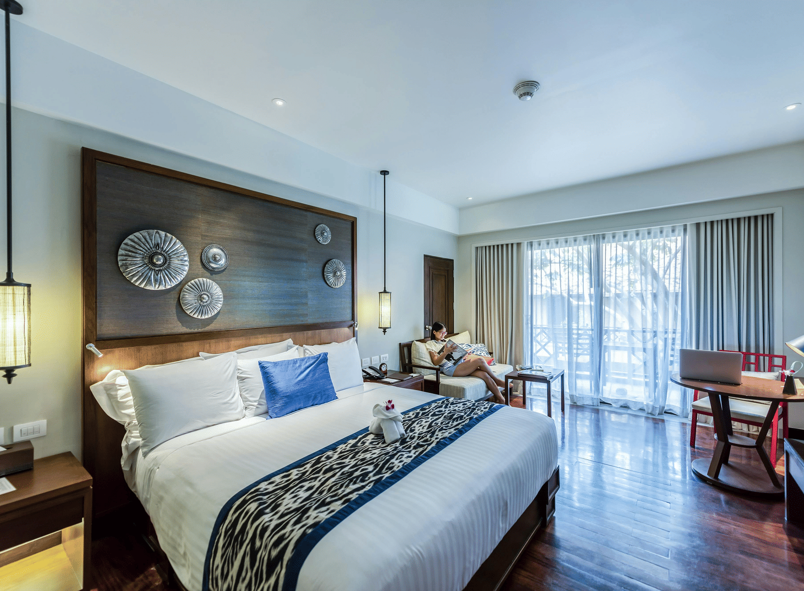 A modern hotel room with a bed, a large decorative headboard, and a seating area by the window.