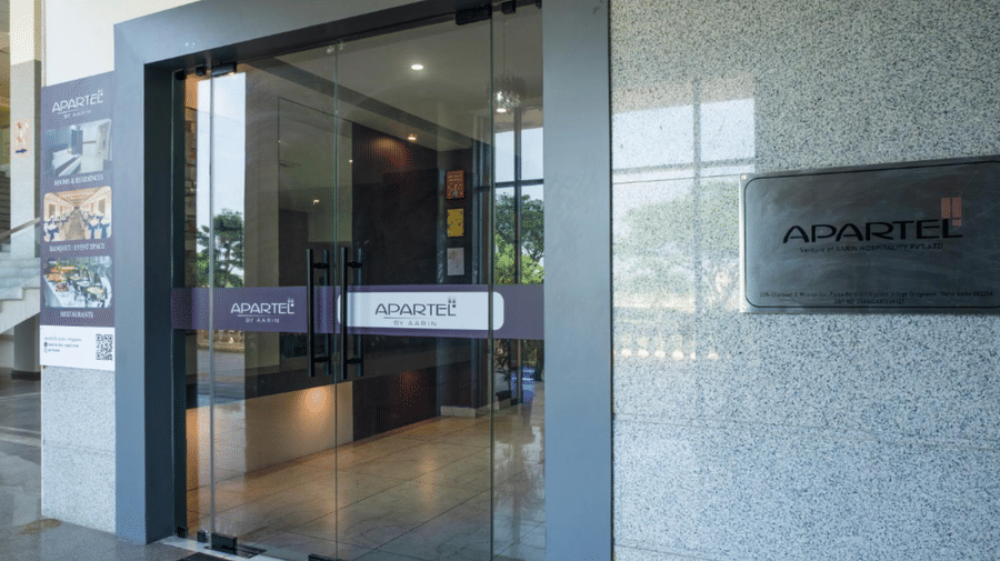 A sleek glass entrance to a modern office or apartment lobby, with grey stone wall panels and subtle branding on a plaque at Apartel by Aarin, Oragadam.