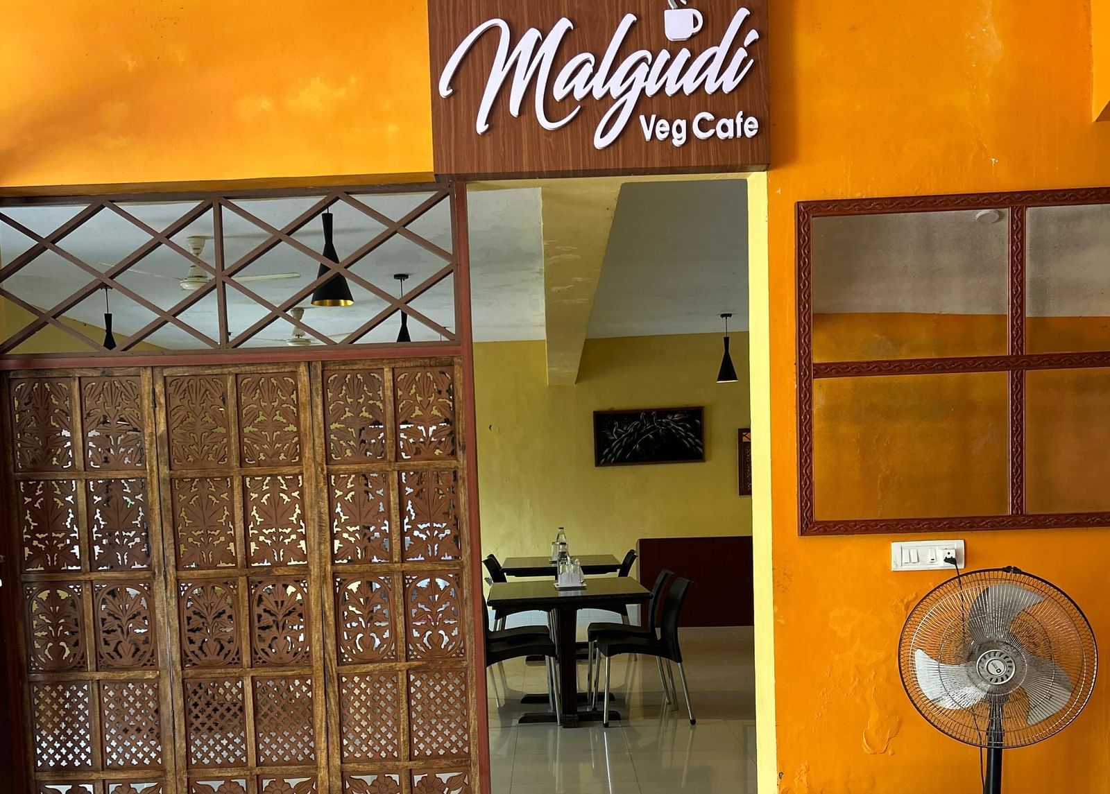 Entrance with wooden partition and yellow wall featuring 'Malgudi' signage outside the dining tables at Paradise Wild Hills Resort, Kollur.