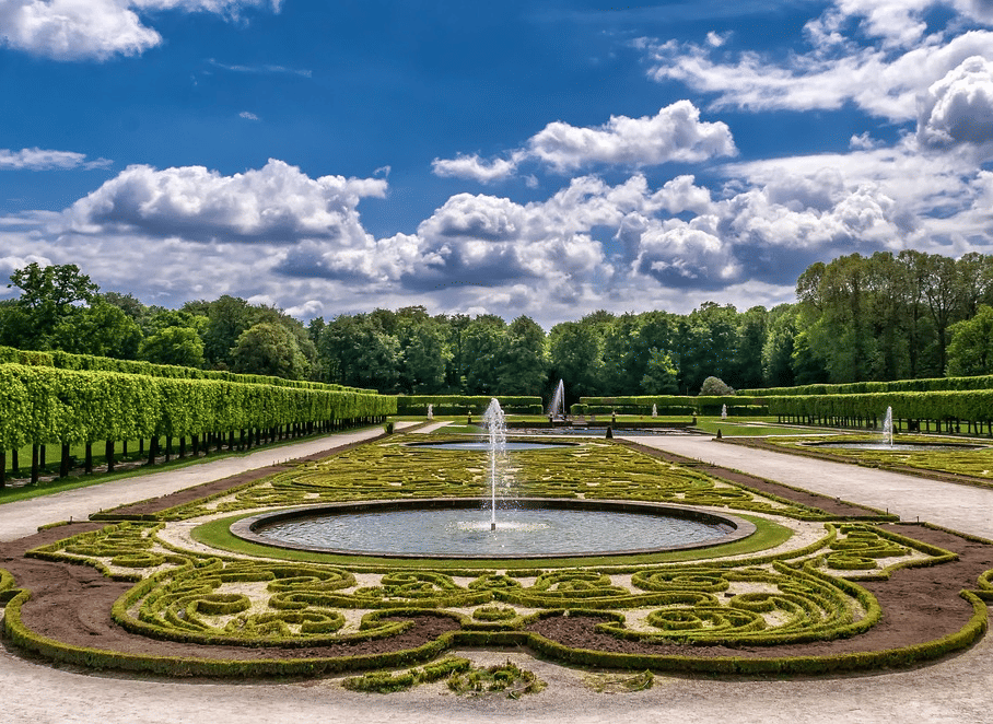 A garden featuring manicured low hedges forming intricate patterns on the ground, centred around a circular fountain and pool