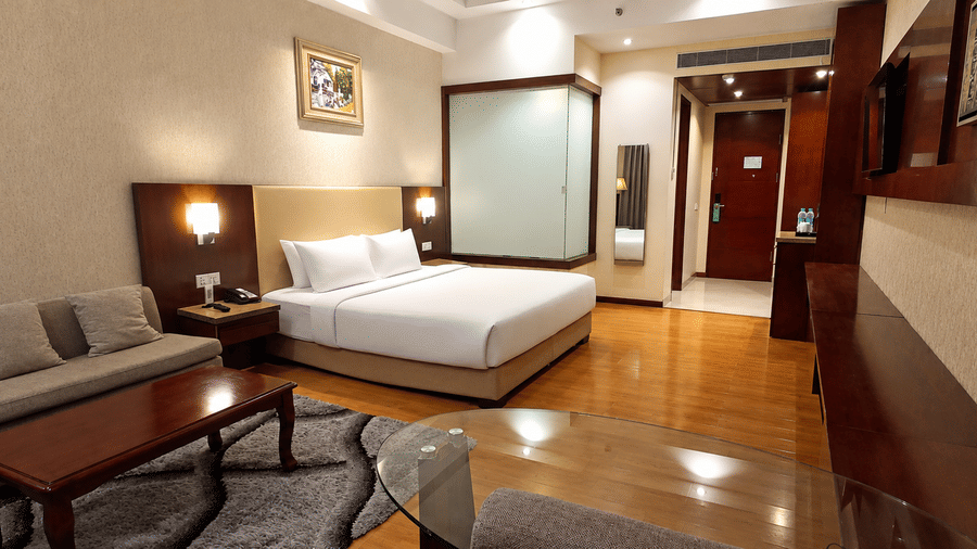 Deluxe room at Golden Tulip Kukas Jaipur with a lounge area featuring a sofa and coffee table in the foreground and a king bed in the background on polished hardwood floors.