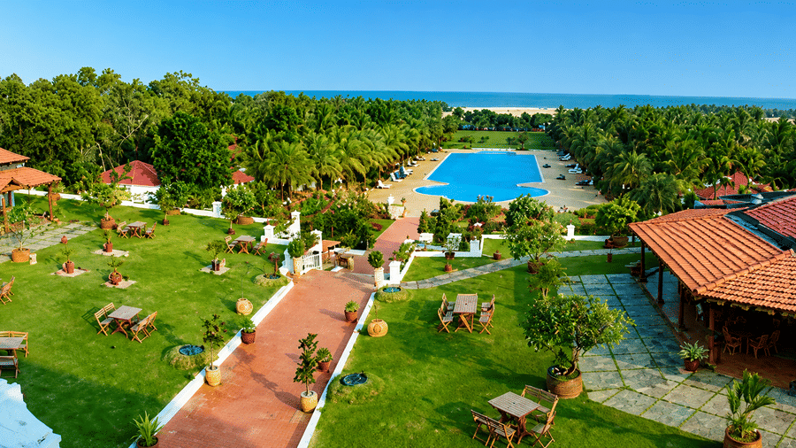 Aerial view of Chariot Beach Resort featuring the pool, lush gardens, red-roofed pavilions, and a stunning ocean backdrop nearby.