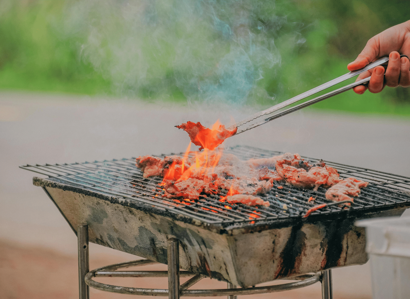 A person's hand uses tongs to turn sizzling meat and vegetables on a barbecue grill.
