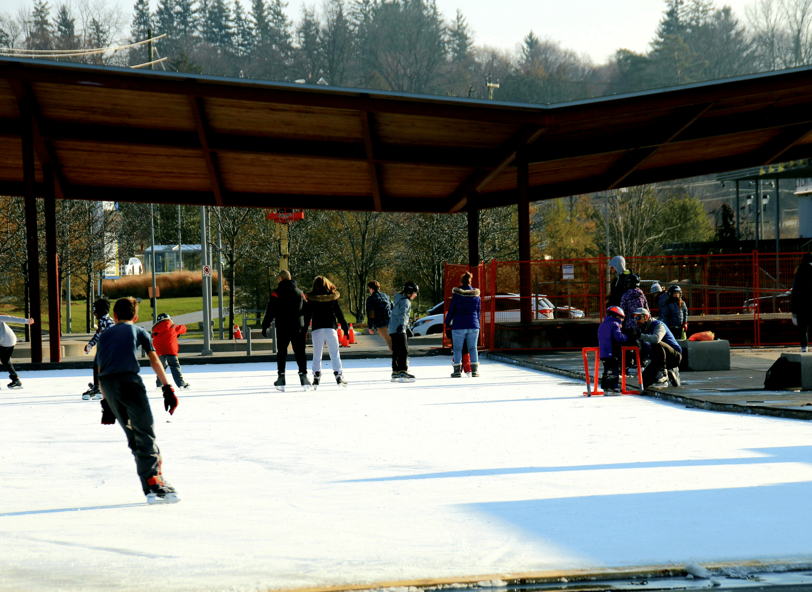 A group of people are ice-skating on a rink under a large, open-sided wooden structure on a sunny day.