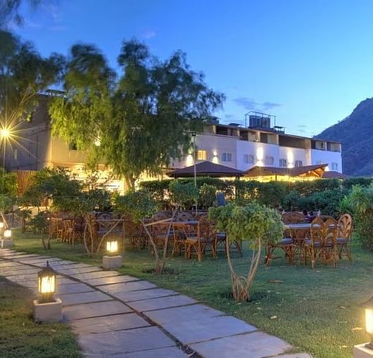 An outdoor scene at dusk where a pathway leads toward a building, which is illuminated with warm lights - Bamboo Saa Resort & Spa, Udaipur