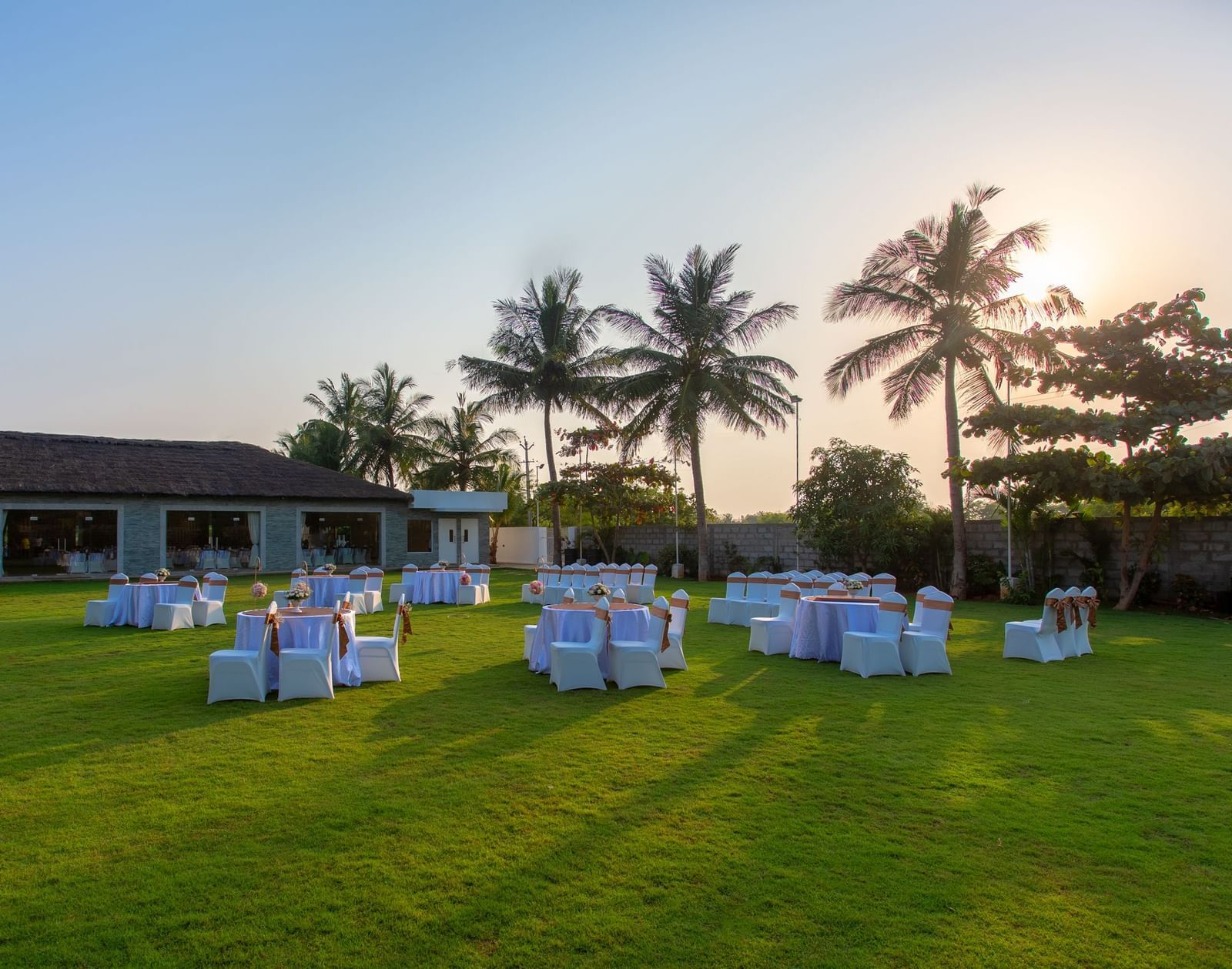 An outdoor event setup on a green lawn with round tables and chairs, and a building with palm trees in the background, under evening light  | Grande Pavillion | Grand Bay Resort and Spa