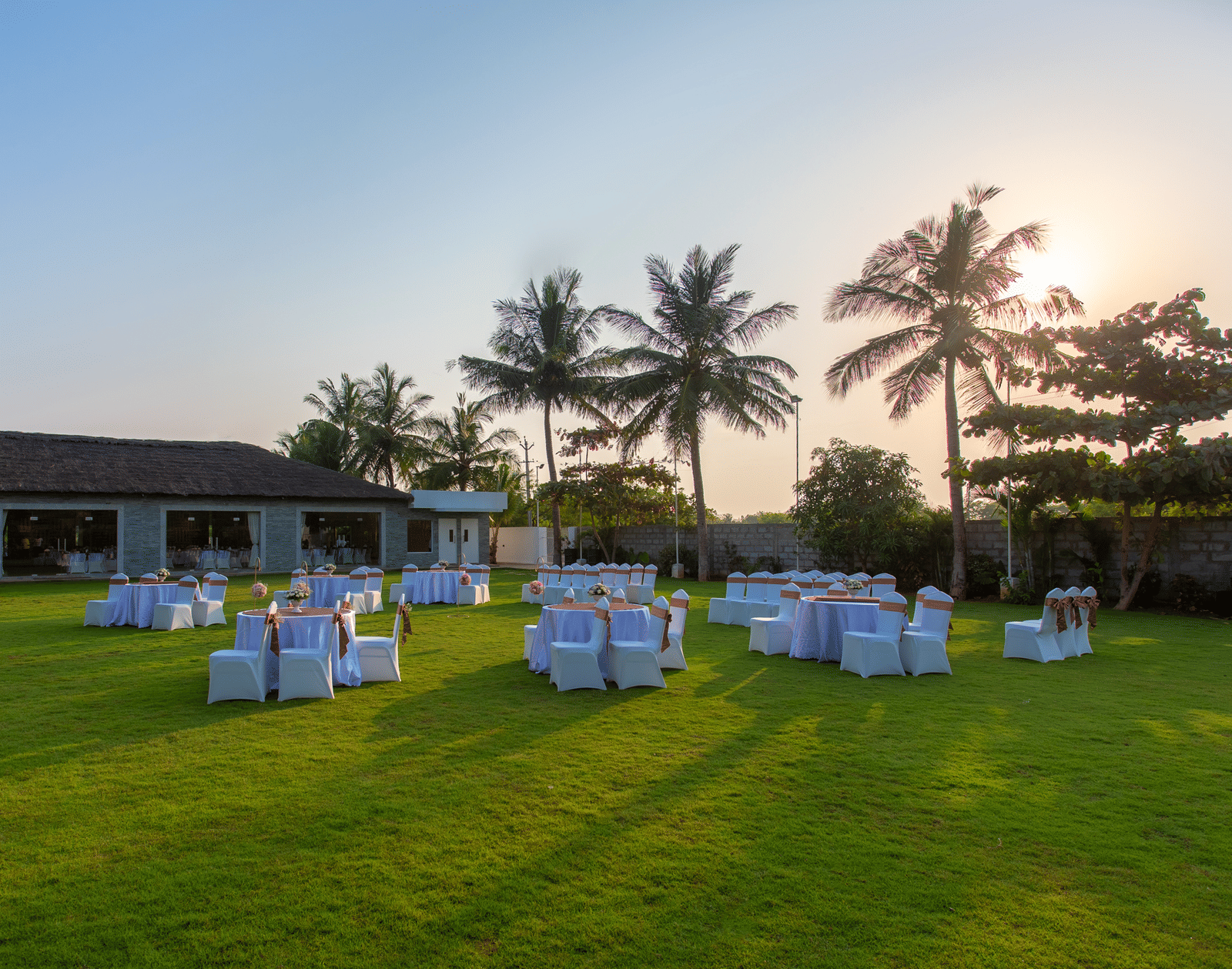 An outdoor event setup on a green lawn with round tables and chairs, and a building with palm trees in the background, under evening light - Grande Bay Resort & Spa, Mamallapuram