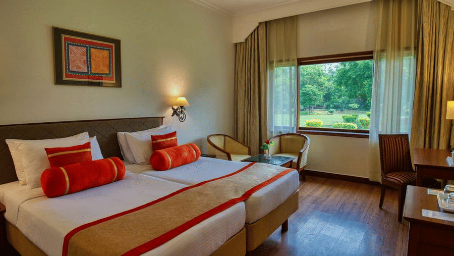 The Deluxe Twin Garden View room at Clarks Khajuraho featuring twin beds pushed together with crisp linens and decorative bolsters along with a wooden work desk, a large window overlooking a lush green garden, and warm accent lighting from bedside lamps.
