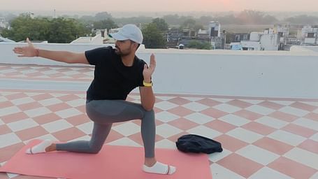 A person performs a yoga pose on a mat on a rooftop with a sunrise or sunset in the background.