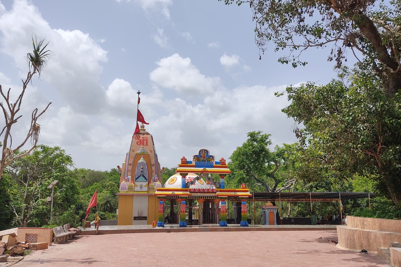 A serene temple painted in bright hues with a tall shikhara and flag, set in an open courtyard surrounded by trees under a partly cloudy sky.