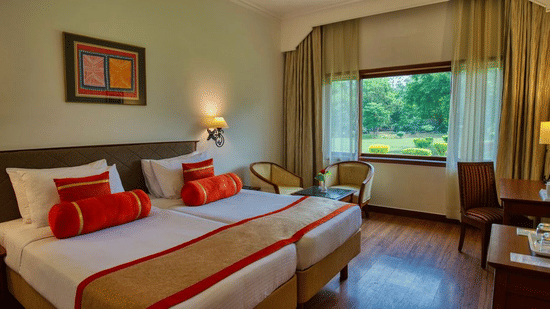 The Deluxe Twin Garden View room at Clarks Khajuraho featuring twin beds pushed together with crisp linens and decorative bolsters along with a wooden work desk, a large window overlooking a lush green garden, and warm accent lighting from bedside lamps.
