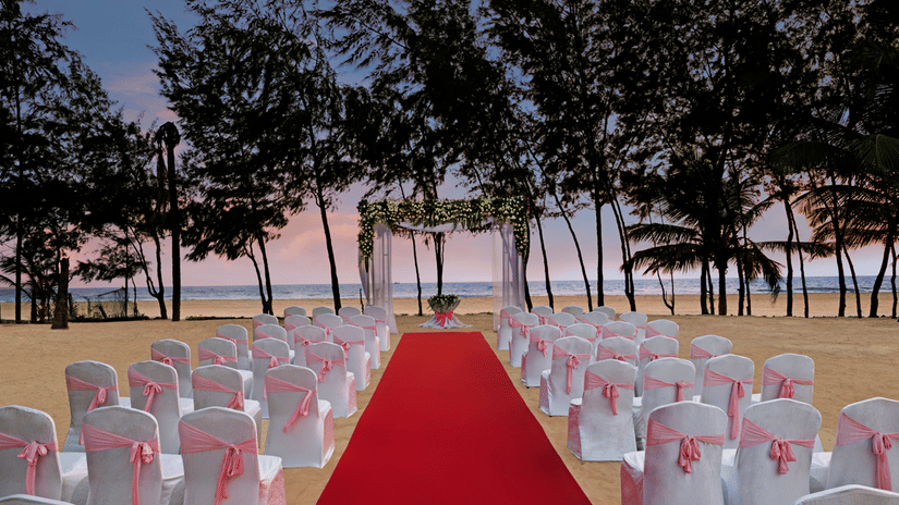 A beach wedding setup at twilight with a red aisle carpet leading to a floral archway, white chairs with pink sashes arranged on the sand, and trees in the background.