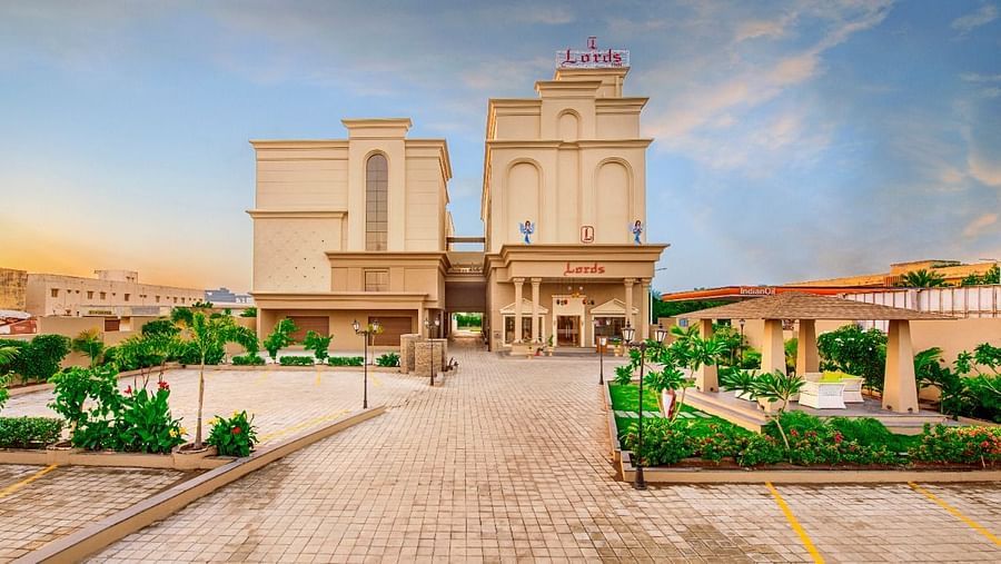 A wide shot of a light-coloured, multi-storey hotel building with a prominent entrance and surrounding greenery, including paved walkways and some parked cars, under a partly cloudy sky at either dawn or dusk at Lords Inn Motikhavdi, Jamnagar.