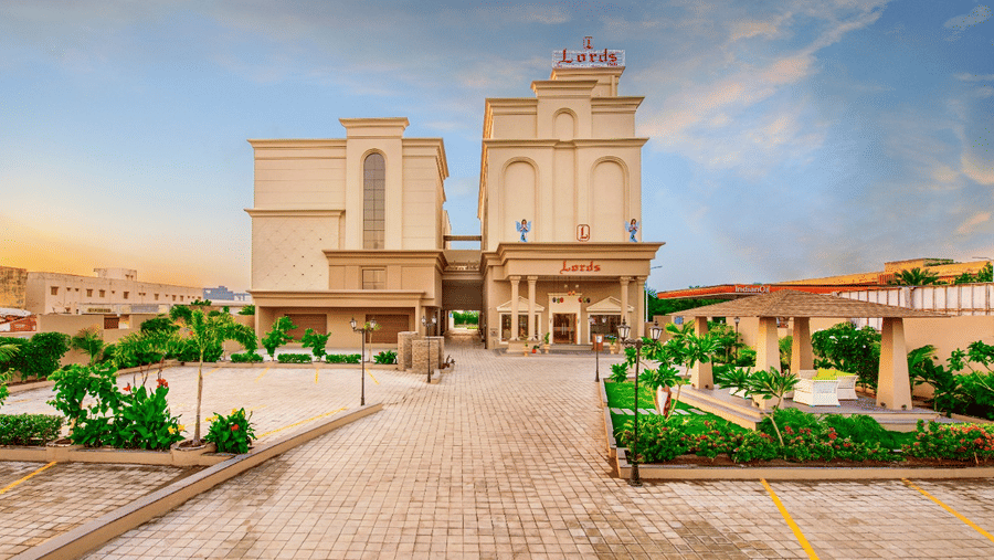 A wide shot of the facade of Lords Inn Jamnagar with a prominent entrance and surrounding greenery, including paved walkways and some parked cars, under a partly cloudy sky 