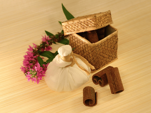 A herbal compress ball (potli) sits next to a woven box and several cinnamon sticks with flowers on a wooden surface.