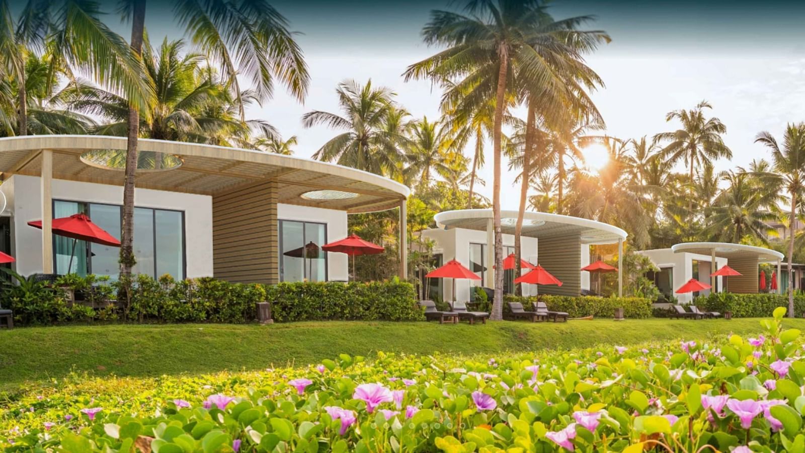 Row of modern white villas with large glass windows, red umbrellas, and sun loungers surrounded by palm trees and flowering greenery at Ramada Resort by Wyndham Khao Lak.